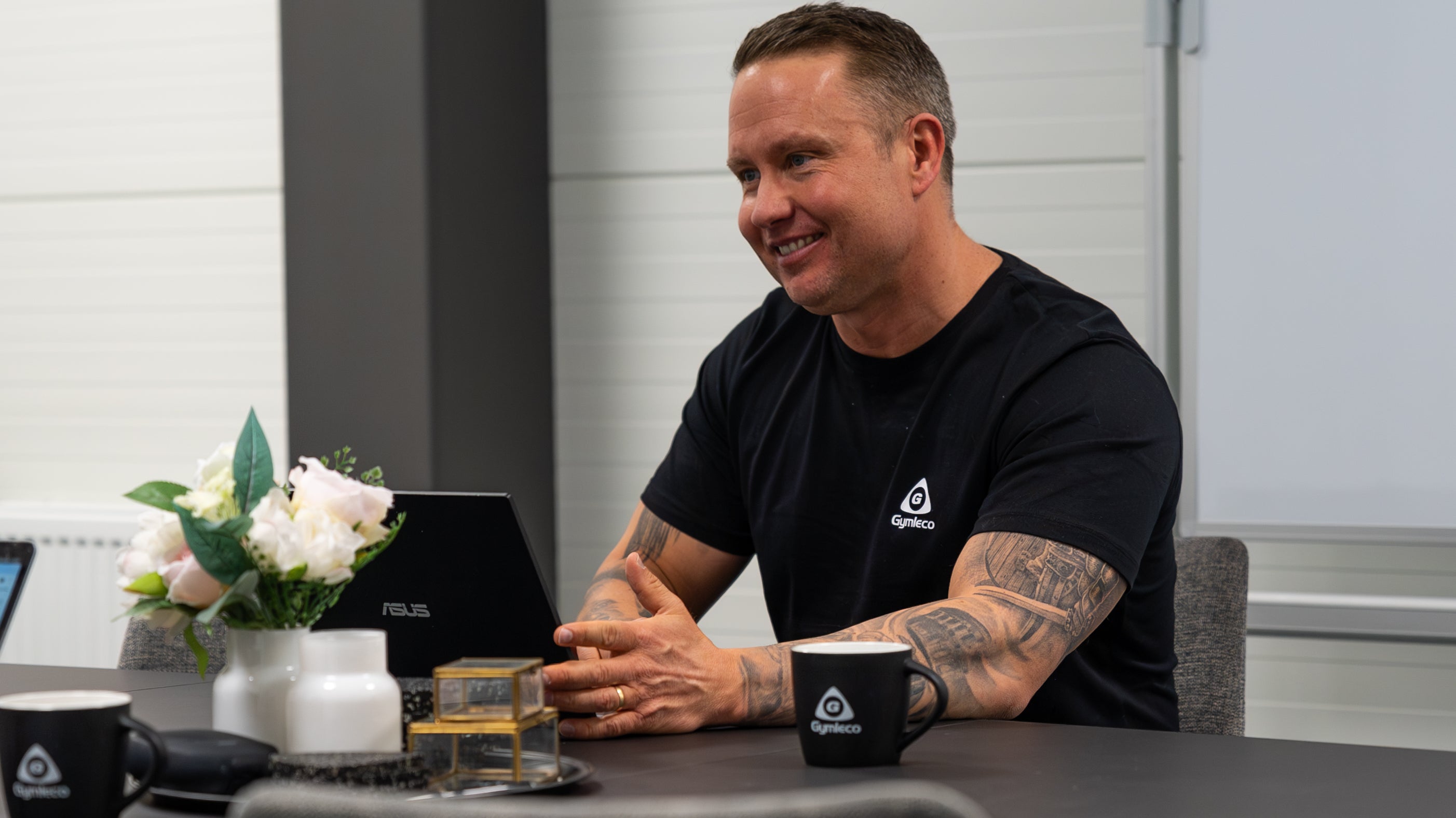 Man sitting at a table with a laptop, mug, and flowers in an office setting.