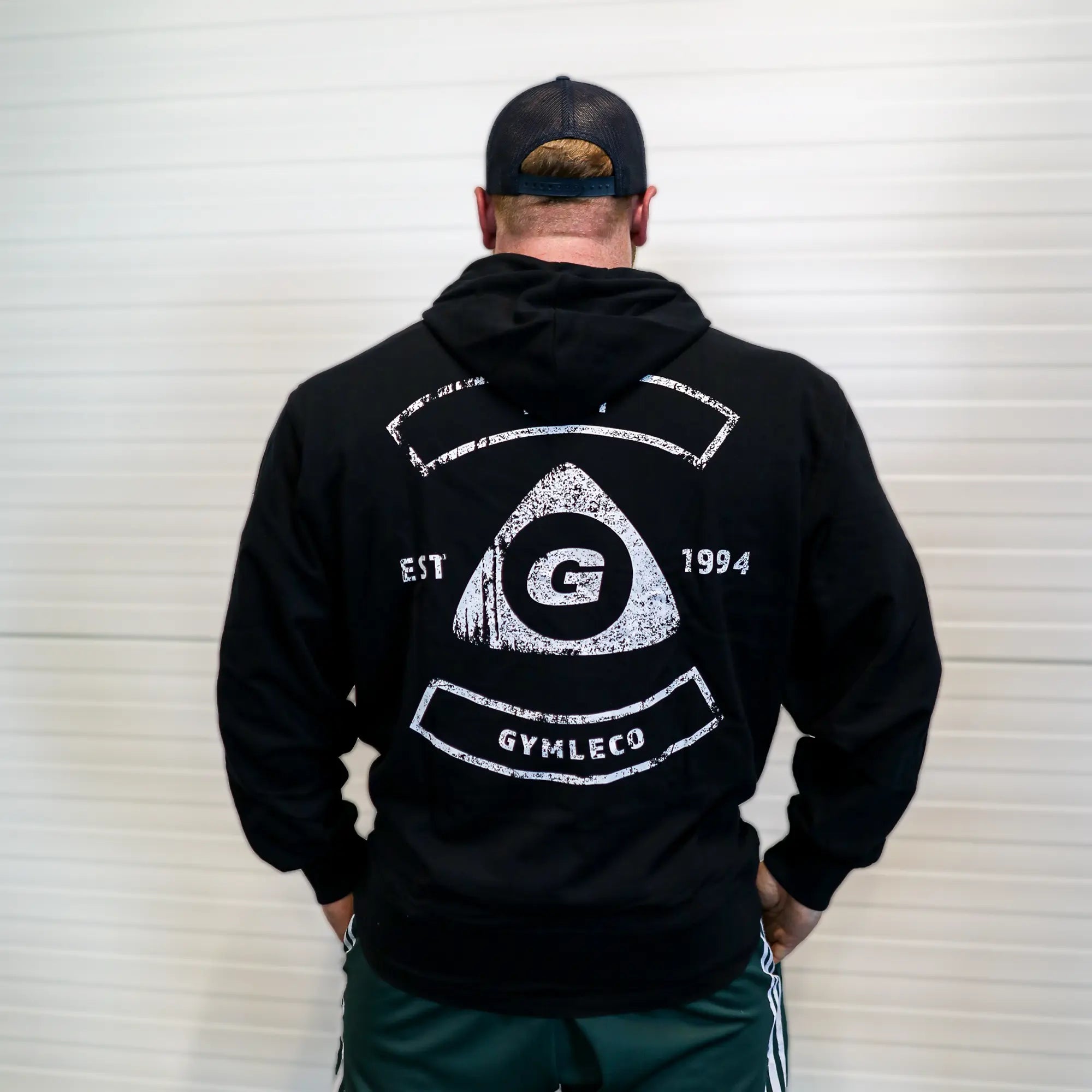 Person wearing a black hoodie with a logo and text on the back, standing against a white wall.