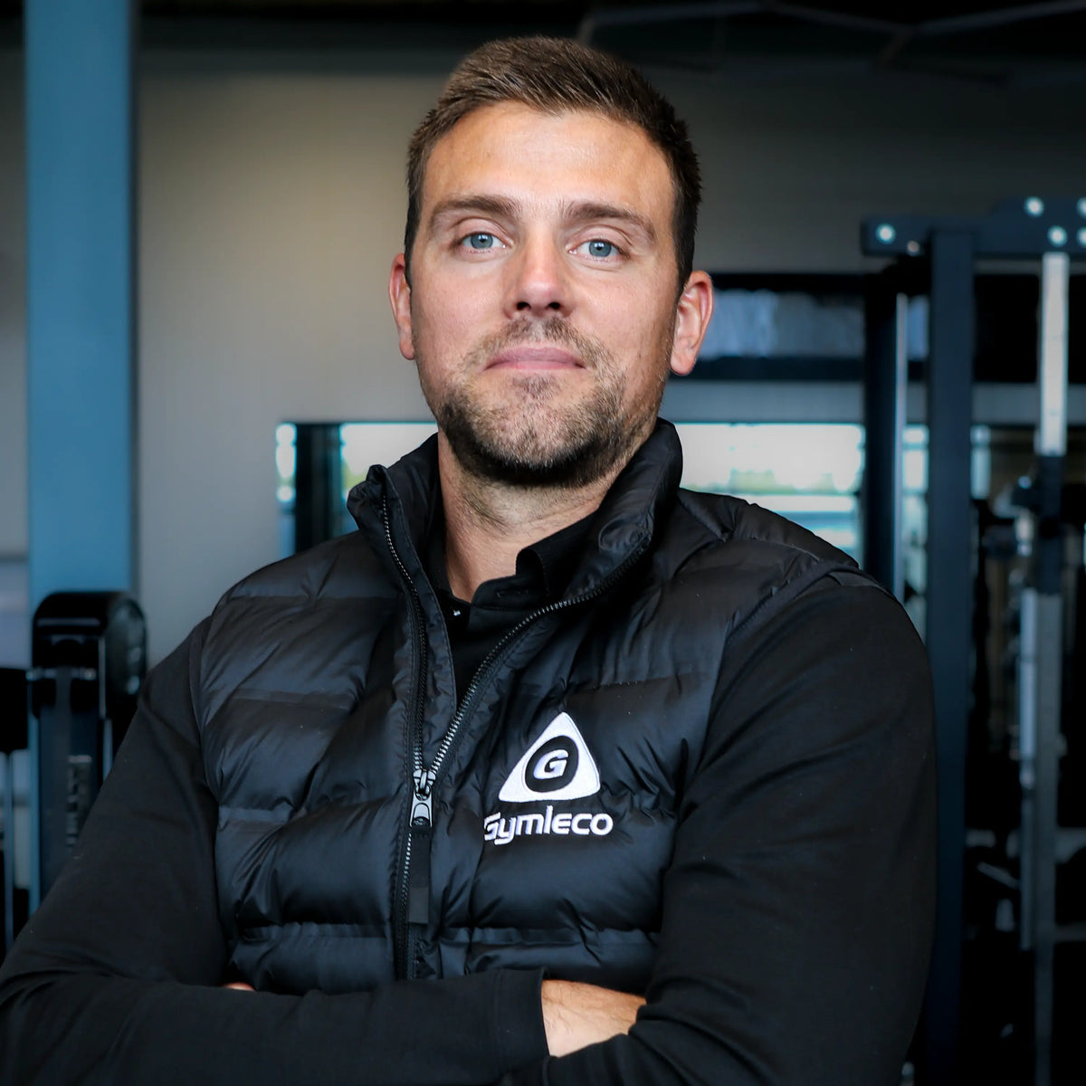 A man with short brown hair and a light beard wearing a black puffer vest with a white 'Gymleco' logo, standing with arms crossed in a gym setting.