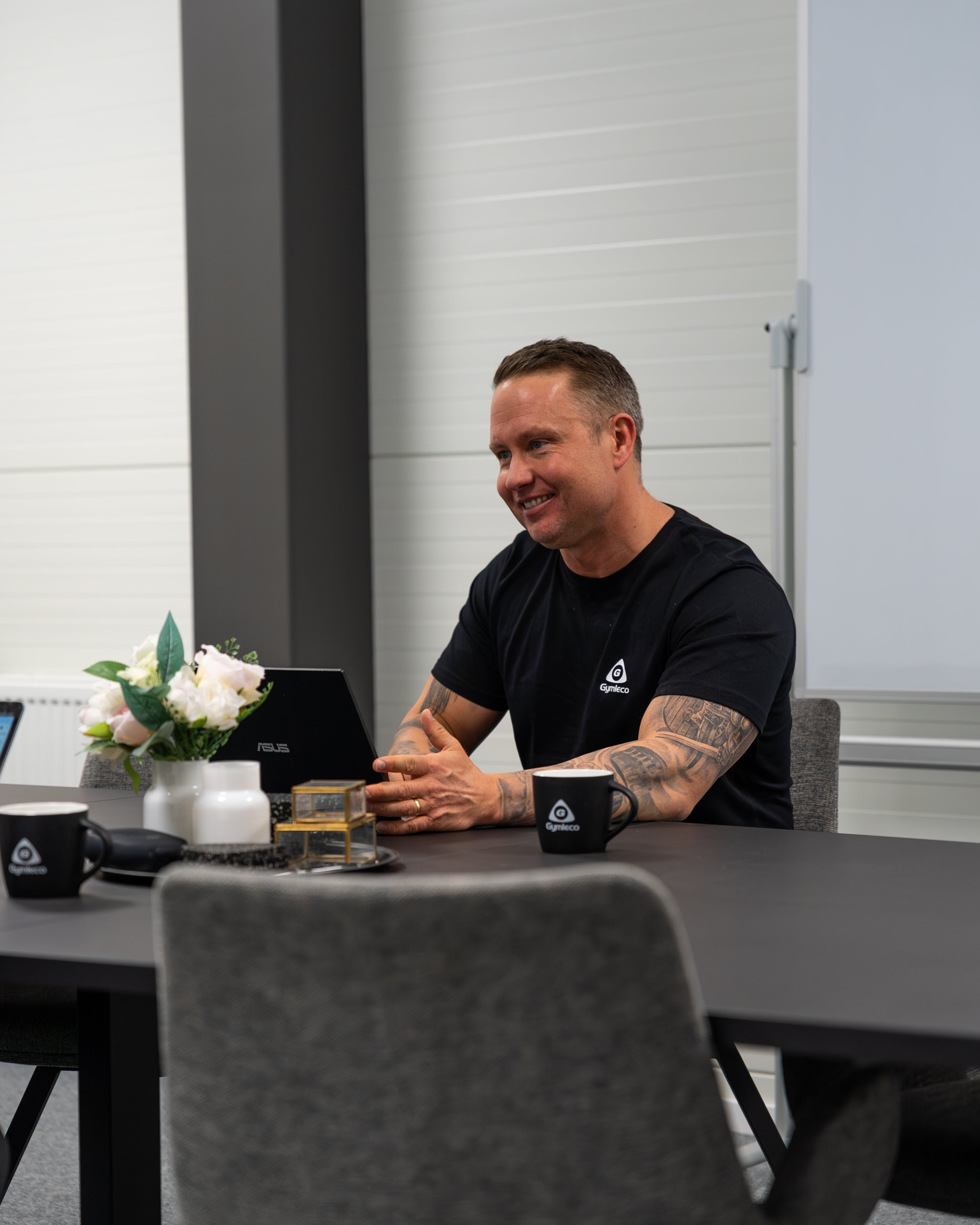 Man sitting at a table with a laptop, mug, and flowers in an office setting.