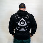 Person wearing a black hoodie with a logo and text on the back, standing against a white wall.