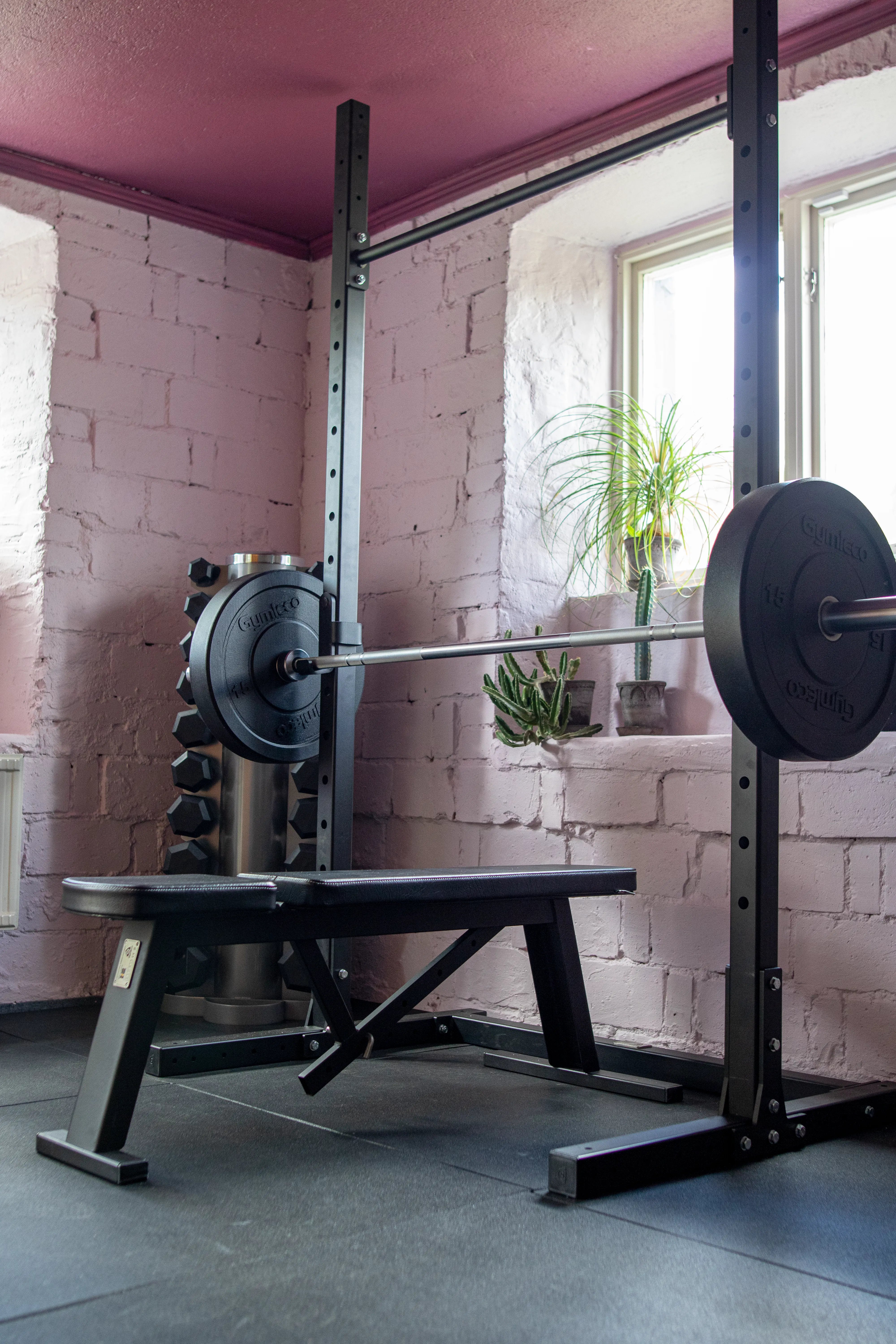 Weightlifting bench with barbell in a room with pink walls and a window.