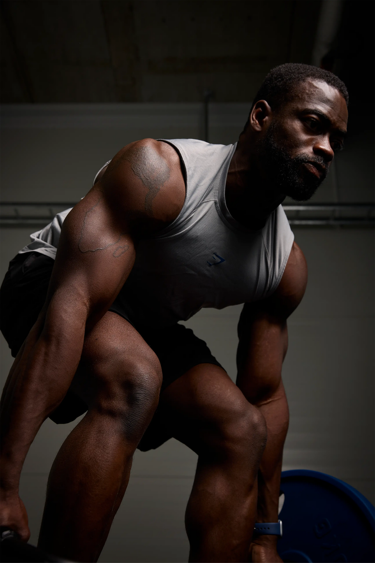 A muscular man in a gym, wearing a light gray sleeveless tank top and black shorts, crouching in a ready stance. The background is a dark, dimly lit gym interior.