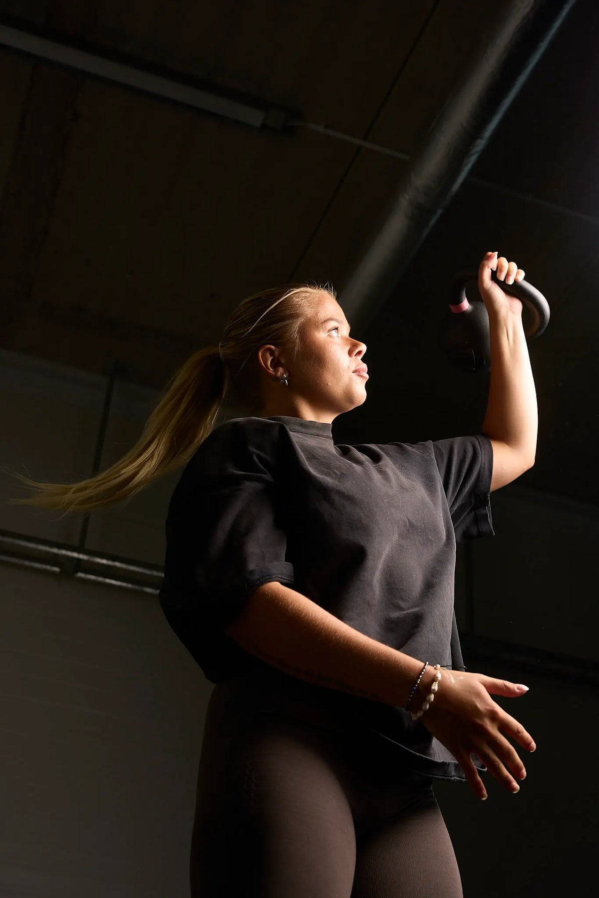 A woman in a gym wearing a black t-shirt and leggings, holding a kettlebell overhead in a dimly lit indoor space.