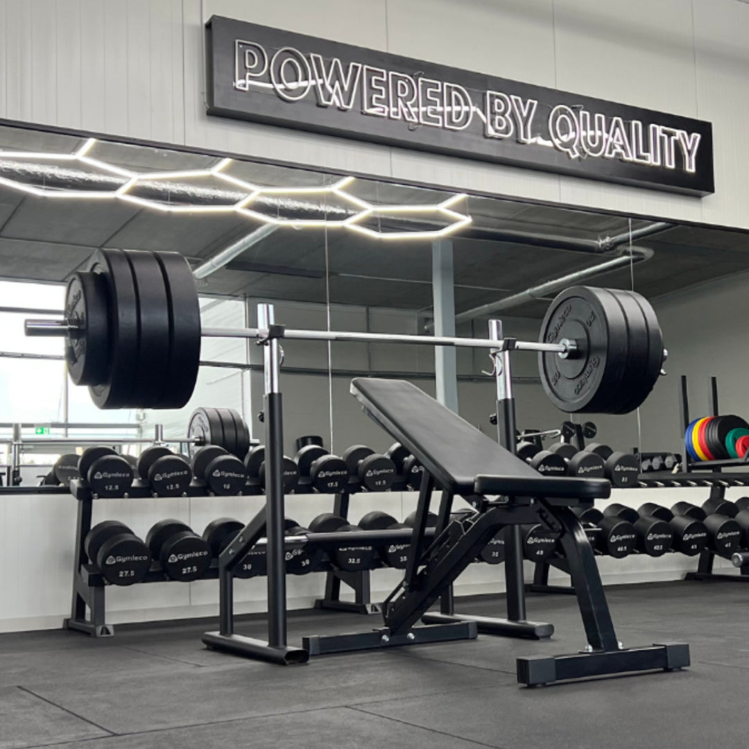 Gym interior with weightlifting equipment and 'Powered by Quality' sign.